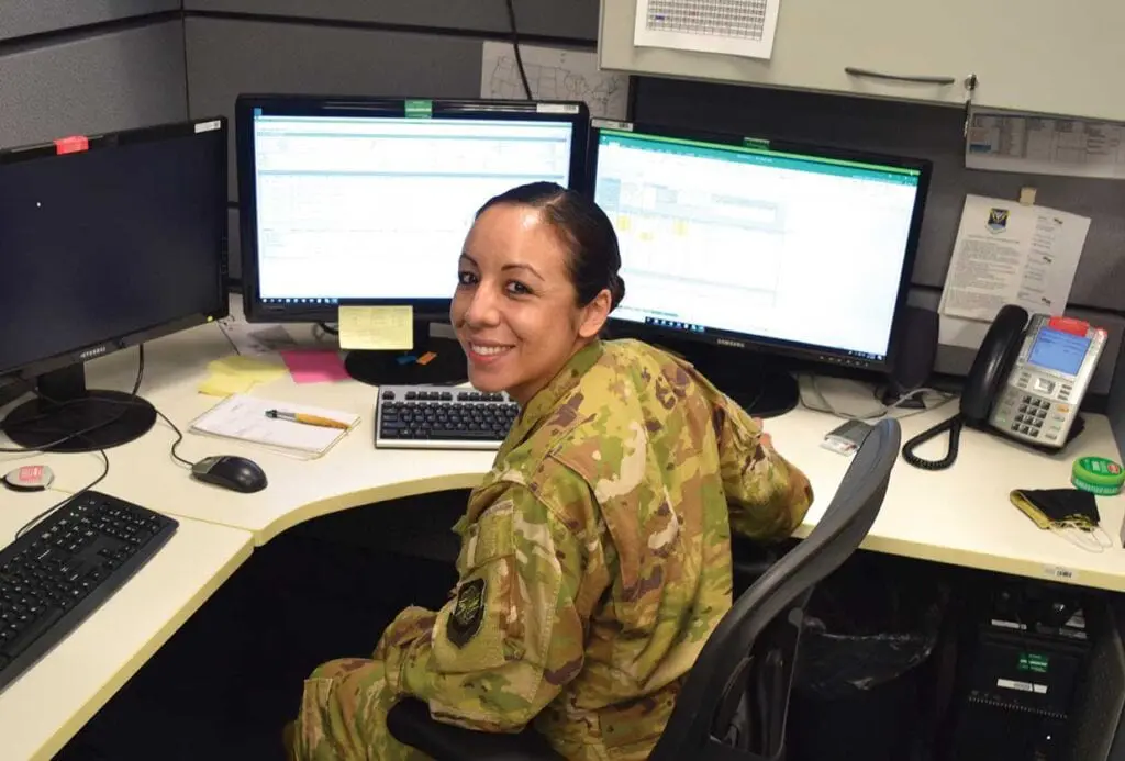 A woman in camouflage sitting at a desk with two computers.