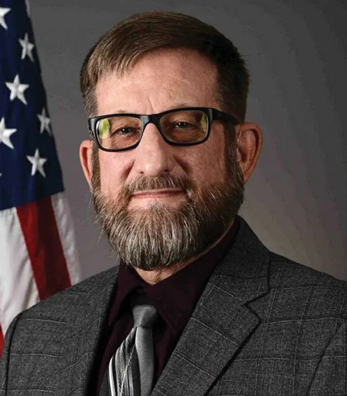A man with a beard and glasses in front of an american flag.