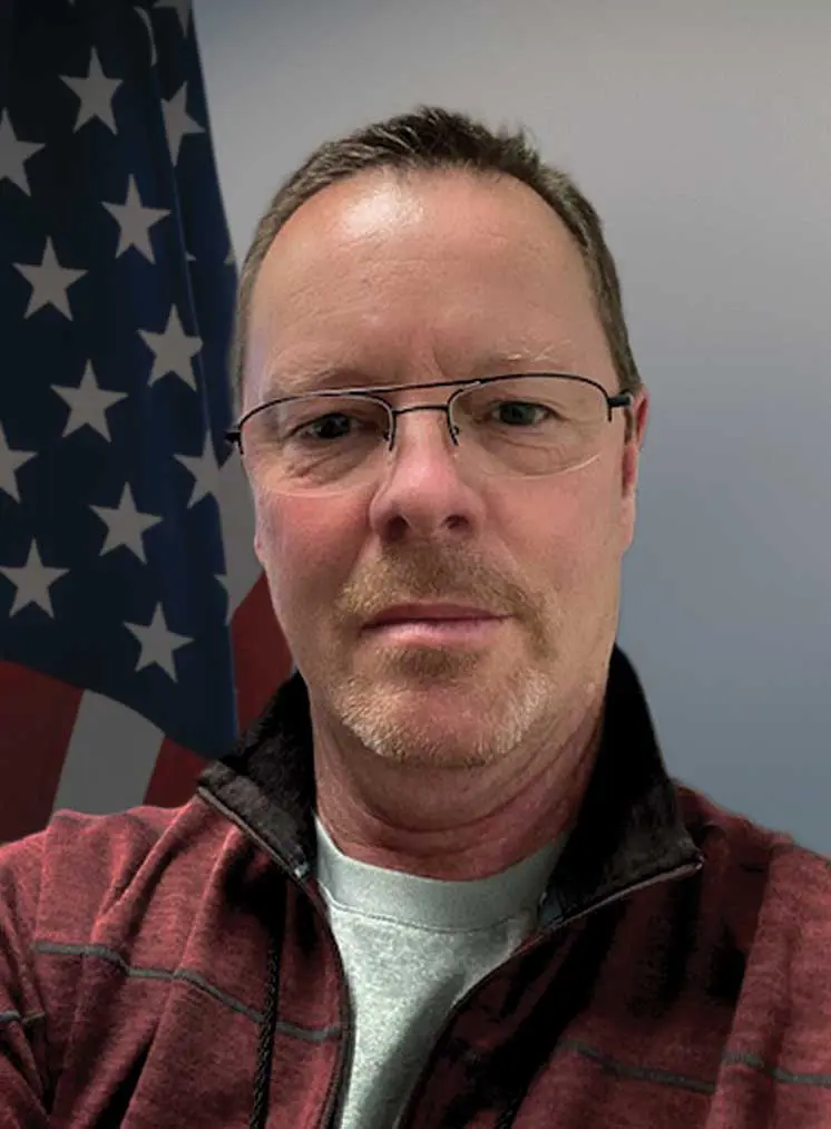 A man with glasses and a red shirt is in front of an american flag.