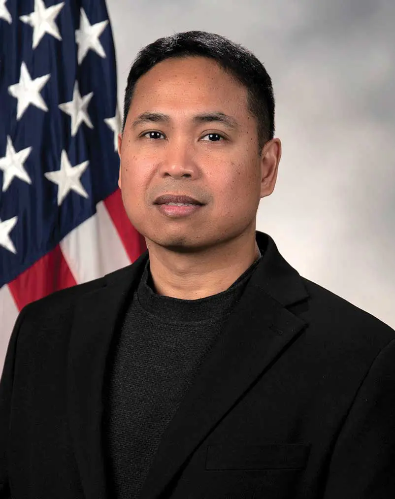 A man in black jacket standing next to an american flag.