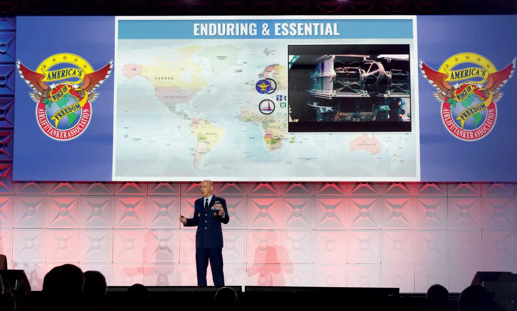 Chief of Staff Gen David Allvin standing on conference stage with a large slide Titled ENDURING & ESSENTIAL with a map and a pop out image of a tank with supplies. The logo America's Wings of Freedom, Airlift/Tanker Association logo on both sides.