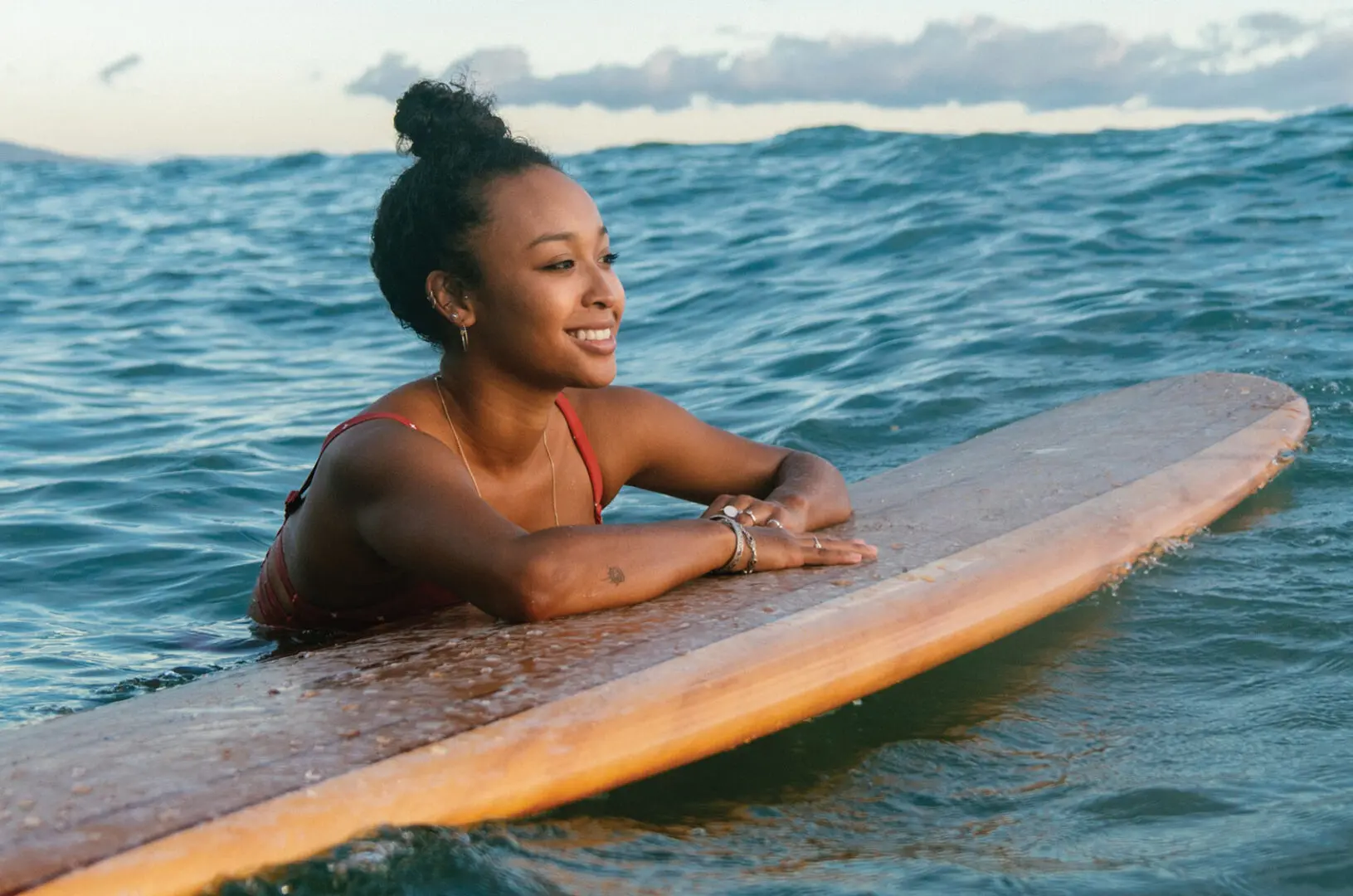 A young woman in a red bathing suit relaxes in the ocean while holding onto a wooden surfboard.