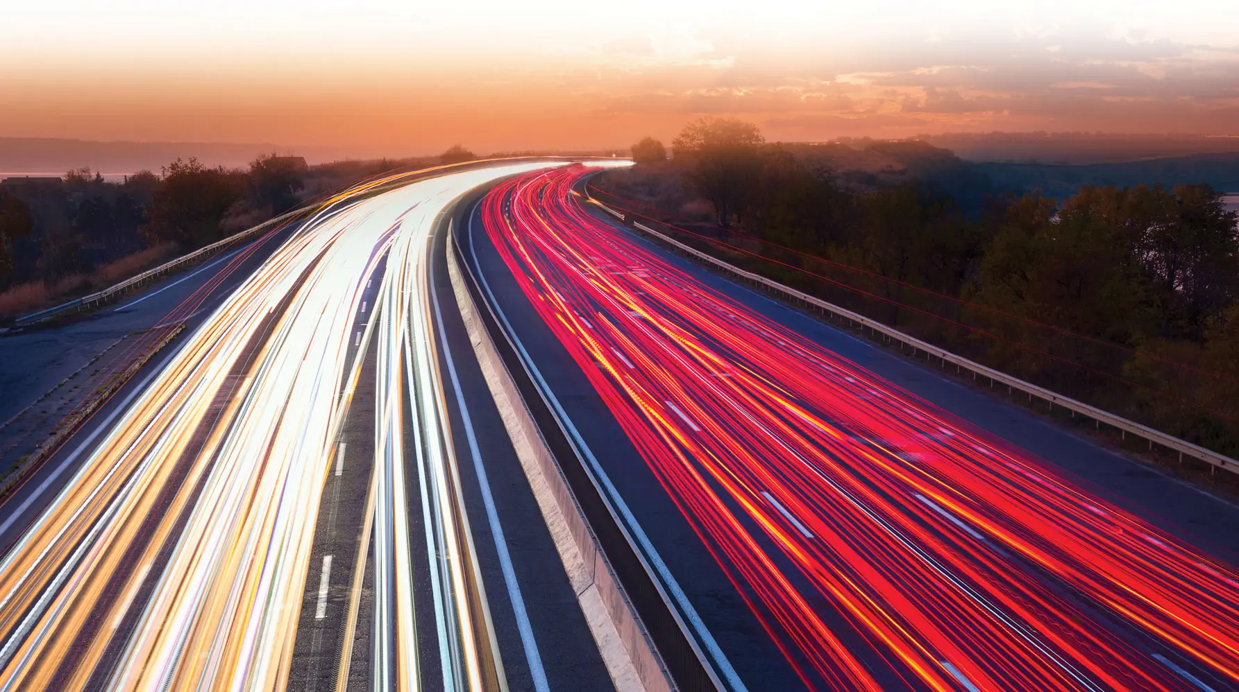 White streaks from car headlights and red streaks from taillights curve around on a highway bordered by trees and trail off into a sunset.