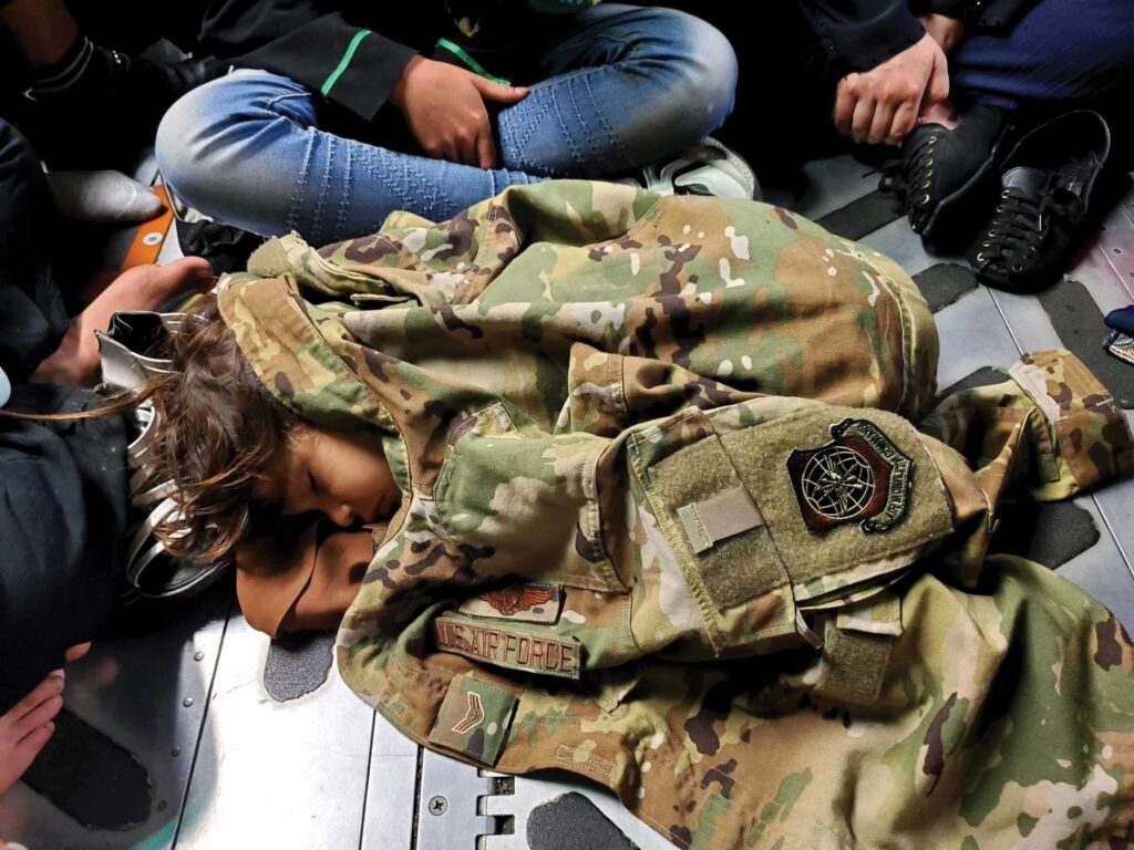 An Afghan child sleeps on the cargo floor of a C-17 Globemaster III, kept warm by the uniform of a C-17 Loadmaster, A1C Nicolas Baron, during an evacuation flight from Kabul, Afghanistan, Aug. 15, 2021. Operating a fleet of Air National Guard, Air Force Reserve, and active duty C-17s, Air Mobility Command, in support of the Department of Defense, moved forces into the theater to facilitate the safe departure and relocation of U.S. citizens, Special Immigration Visa recipients, and vulnerable Afghan populations from Afghanistan. USAF photo by Capt Mark Lawson