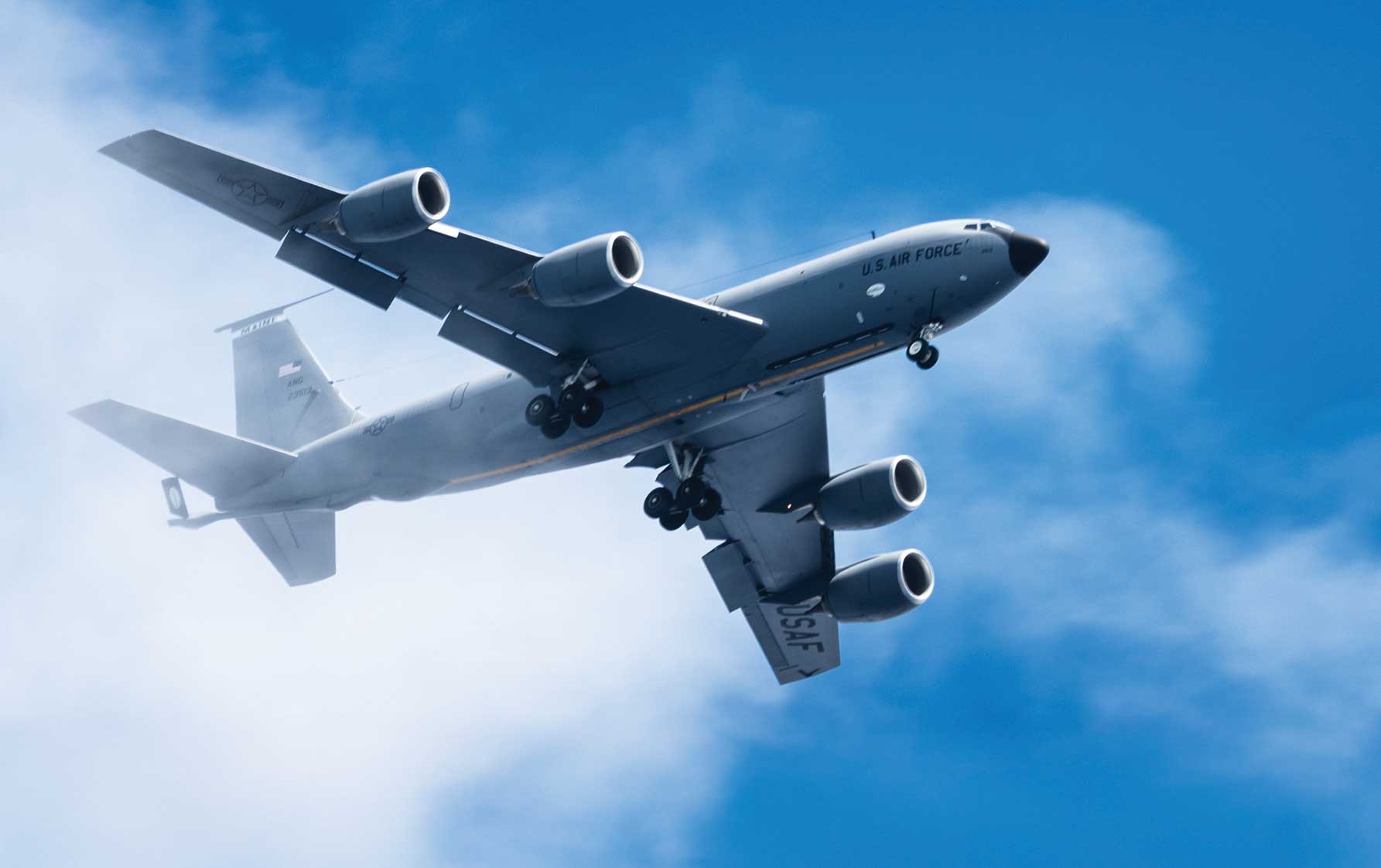 A KC-135 Stratotanker aircraft flying out of clouds under a blue sky.