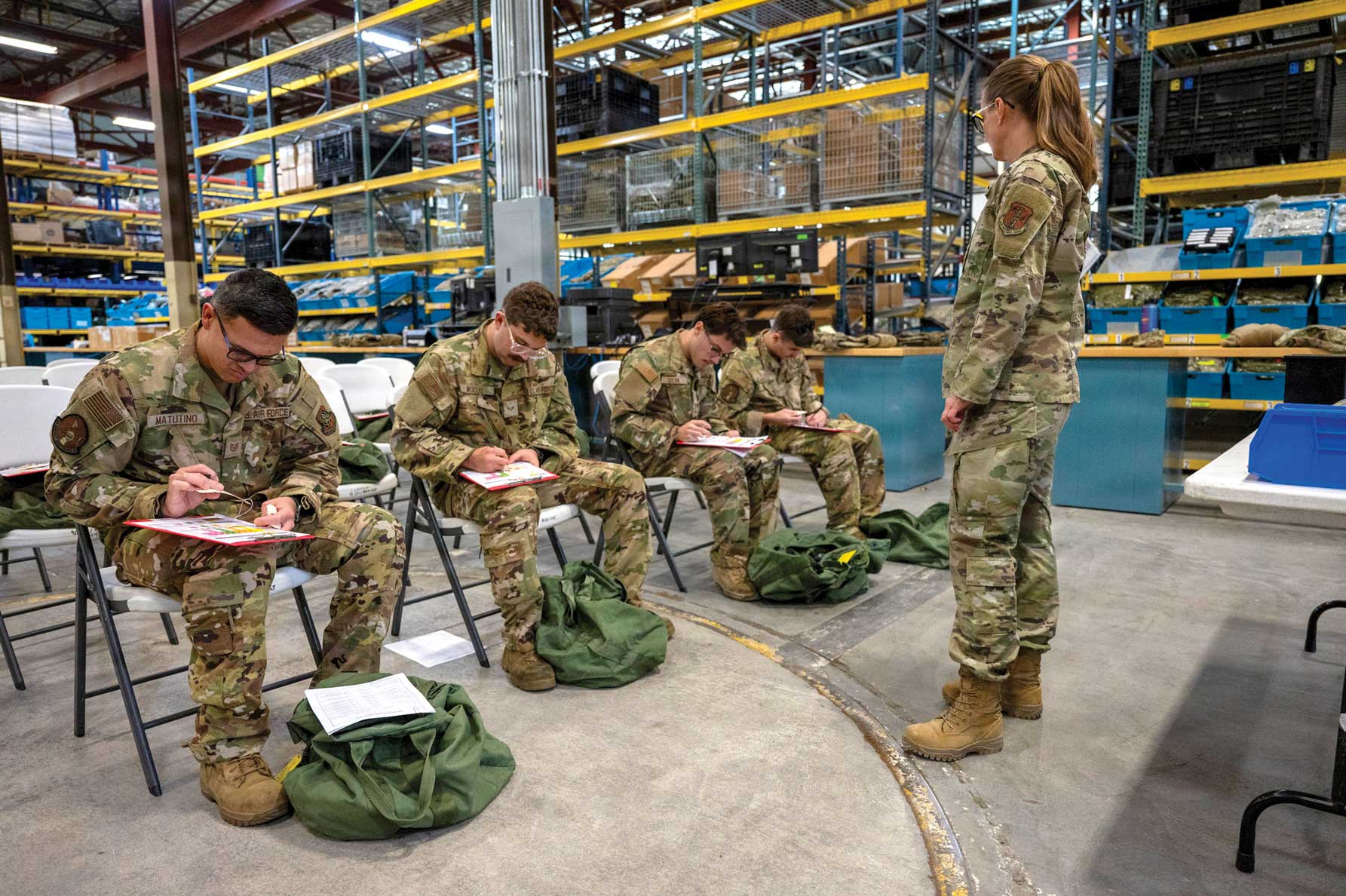 Four Airmen sit in chairs filling out paperwork while a female Airman looks on.