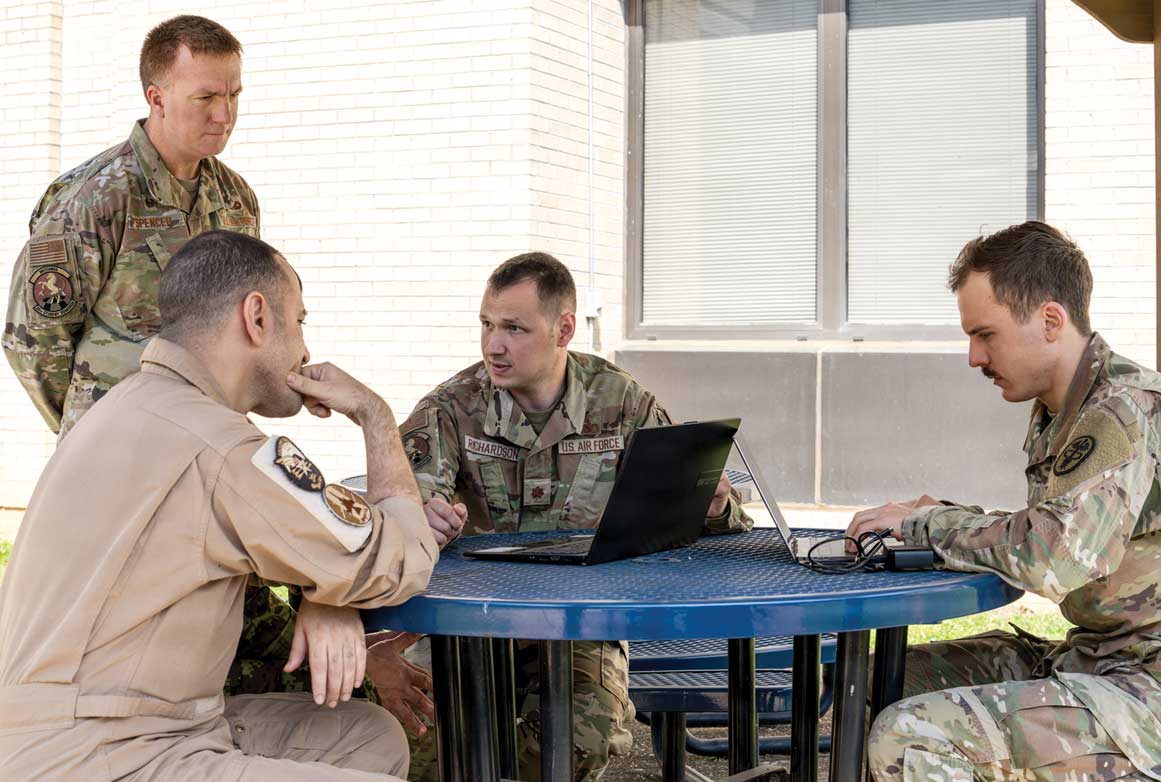 An Airman stands near a table where three other Airmen are sitting and looking at laptop computers.