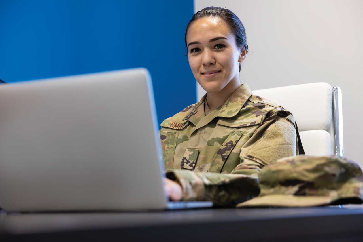 A woman in a military uniform is sitting at a desk using a laptop.