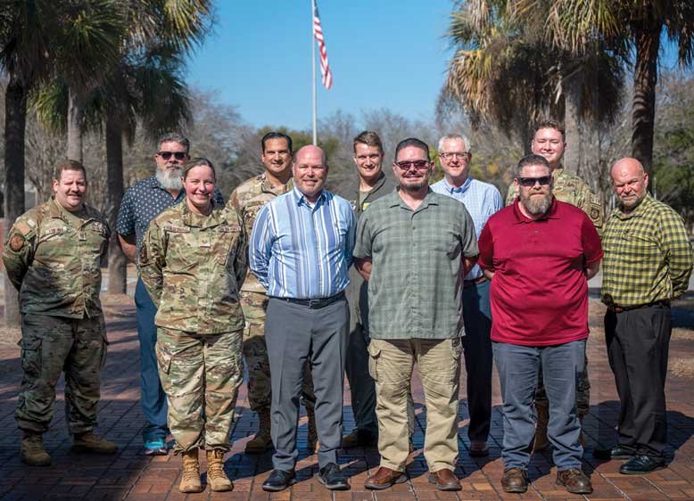 Group of men, some in casual clothes and one in military uniform, standing outdoors on a sunny day.