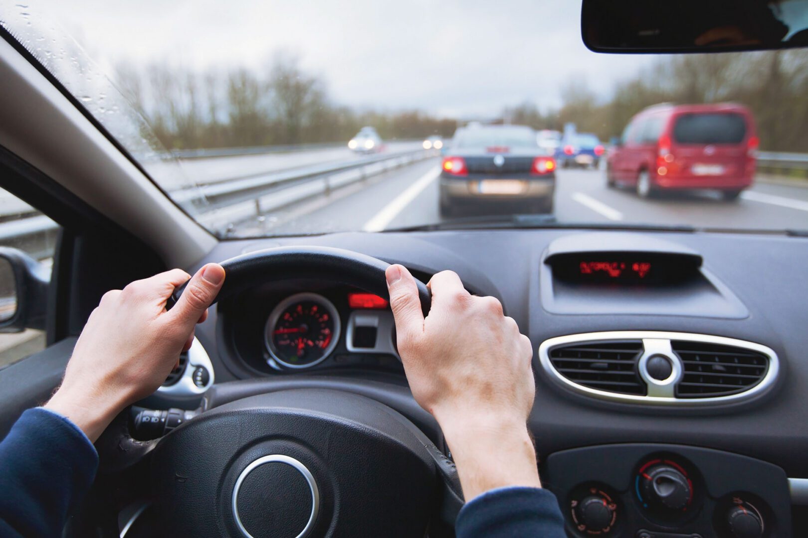 Hands on a steering wheel and interior of a car, with the view through the front windshield of traffic on a highway.