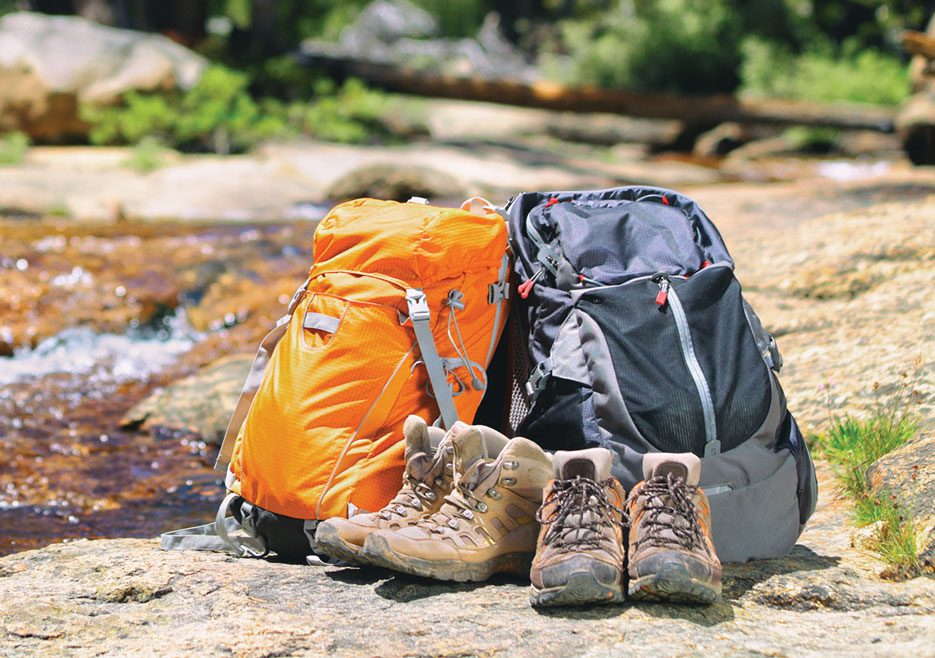 Two backpacks and two pairs of hiking shoes sitting on rocky ground next to a stream.