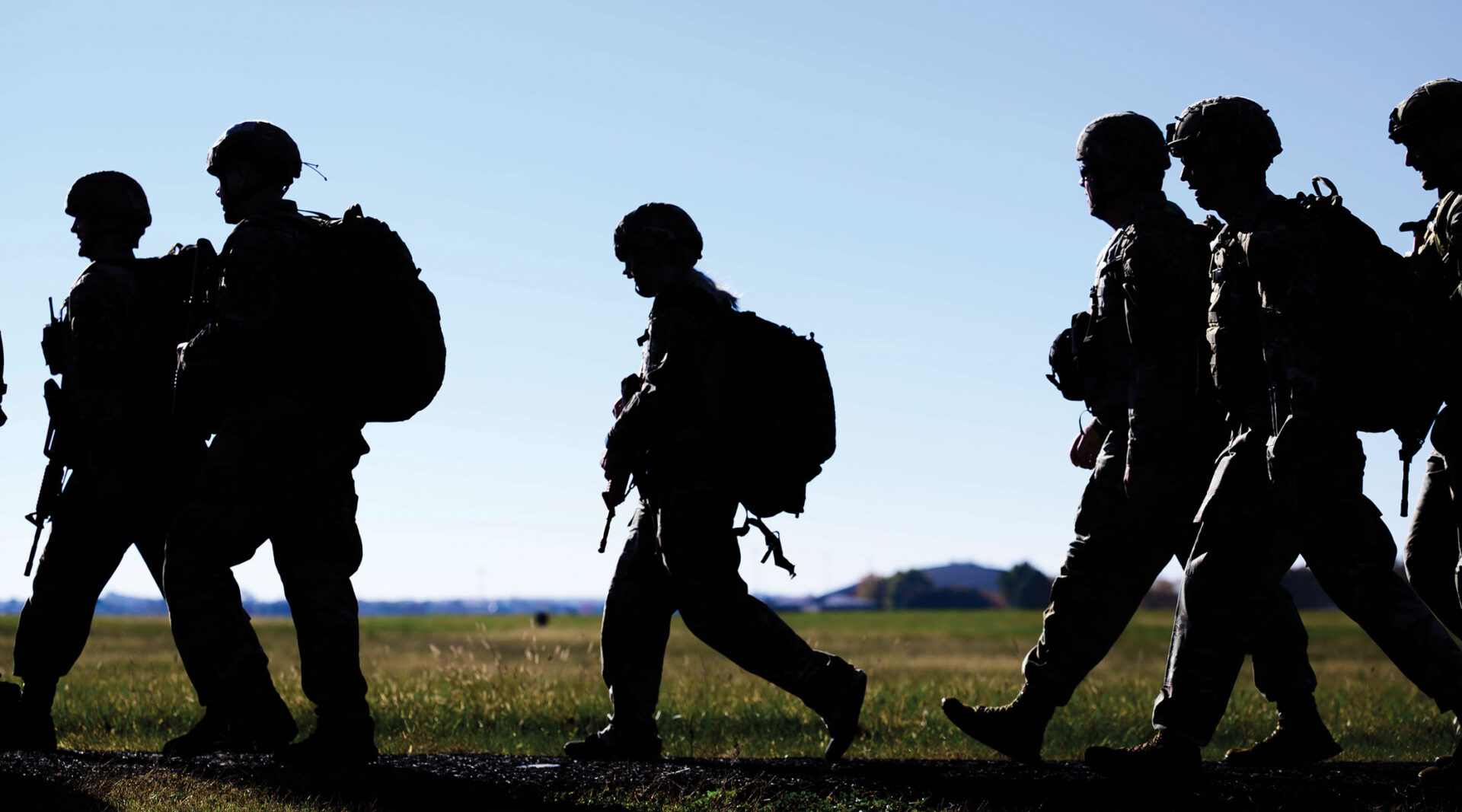 Silhouettes of six Airmen in full gear, walking on a path by a field.