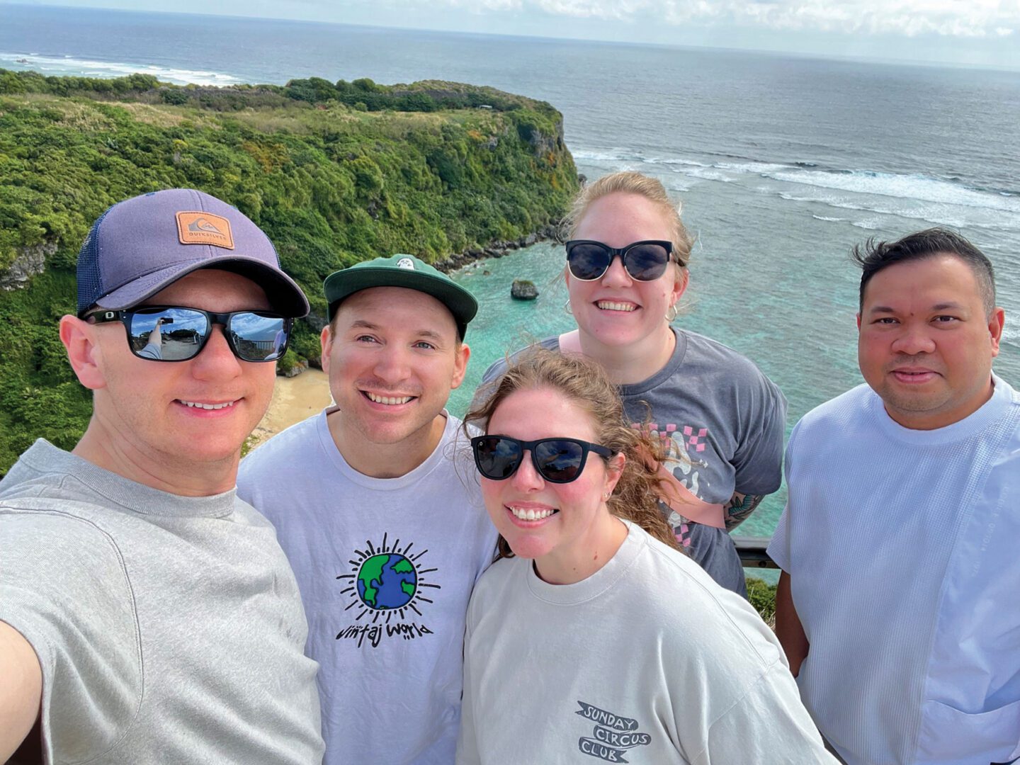 Three men and two women pose for a photo in front of a green hill and a body of water.