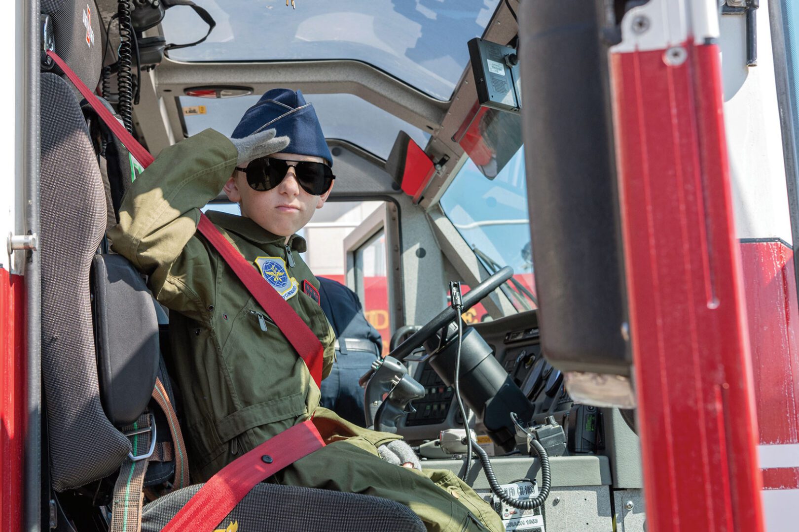 A young child wearing a pilot uniform and dark sunglasses sits in an aircraft, giving a salute.
