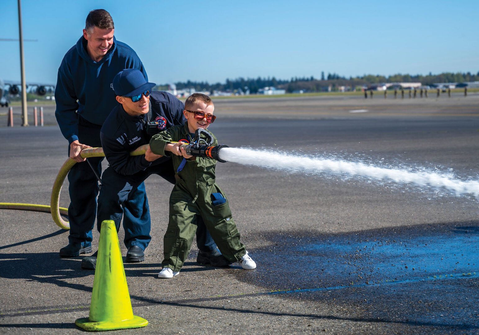 An Airman helps a young child hold a firehose spraying water.