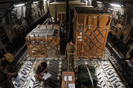 An Airman kneels in front of cargo in an airplane, looking at paperwork, while another Airman pulls a strap over boxes.