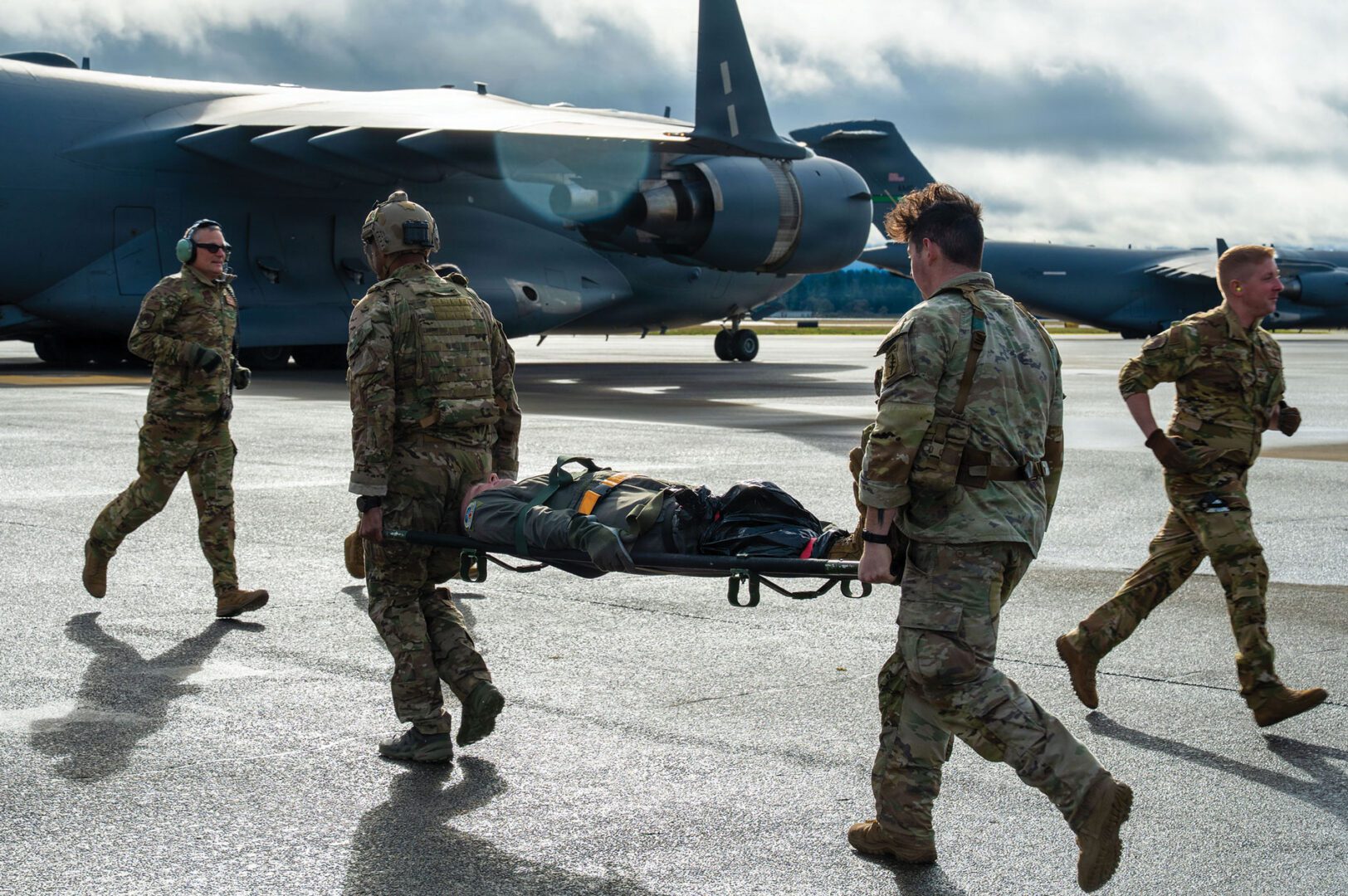 Two Airman carry a person on a stretcher towards an aircraft on a flightline, while two other Airman run the other way.