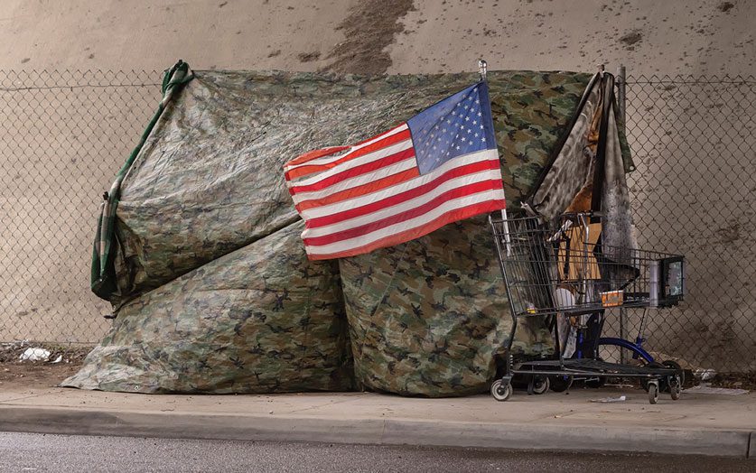 A camo-colored tarp fashioned into a tent and a shopping cart with an American flag attached to it sit in front of a chain-link fence in an underpass.